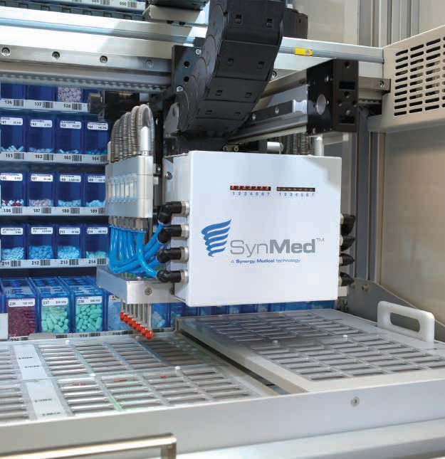 Close view of the SynMed XF robotic arm positioned over a blister card tray, with medication canisters in the background.